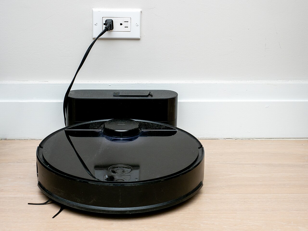 Robot aspirador negro cargando en su base junto a la pared, con espacio libre alrededor